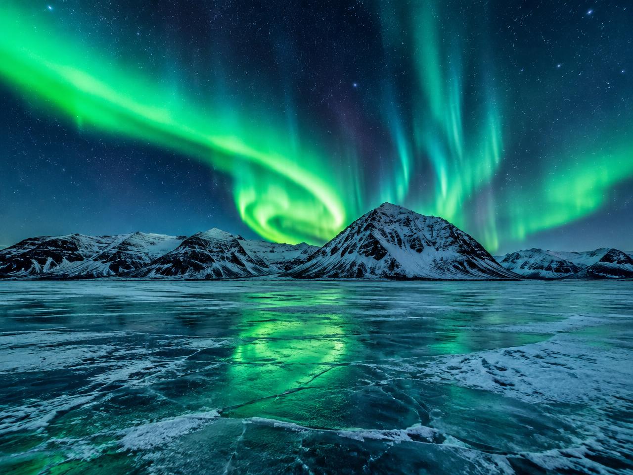 Green northern lights swirling above snowy mountains and a frozen lake in Iceland